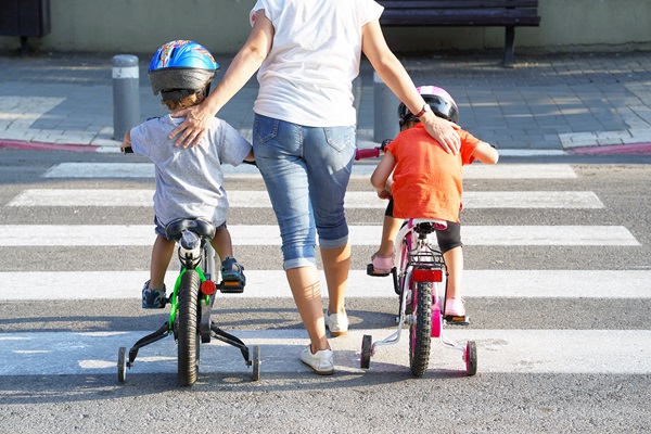 Parent guiding two young children on bicycles with training wheels across a crosswalk.